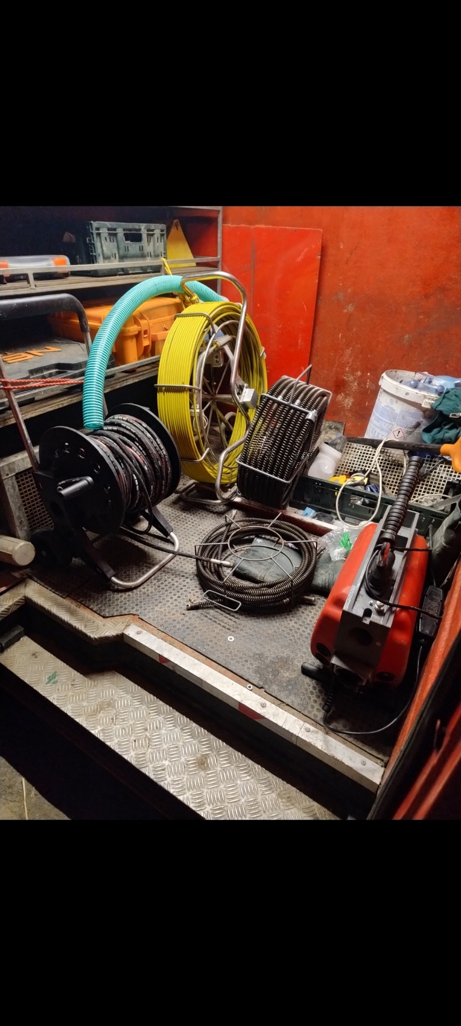 Wnętrze pojazdu serwisowego z różnymi narzędziami do czyszczenia i inspekcji rur, w tym zwinięte przewody, elektryczna maszyna do przepychania rur z czerwonym korpusem i żółty wąż. Na podłodze...