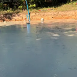 Świeżo wylana, gładka płyta fundamentowa z wystającymi rurami instalacyjnymi, otoczona wykopem i ziemią, widoczne narzędzia budowlane w tle.