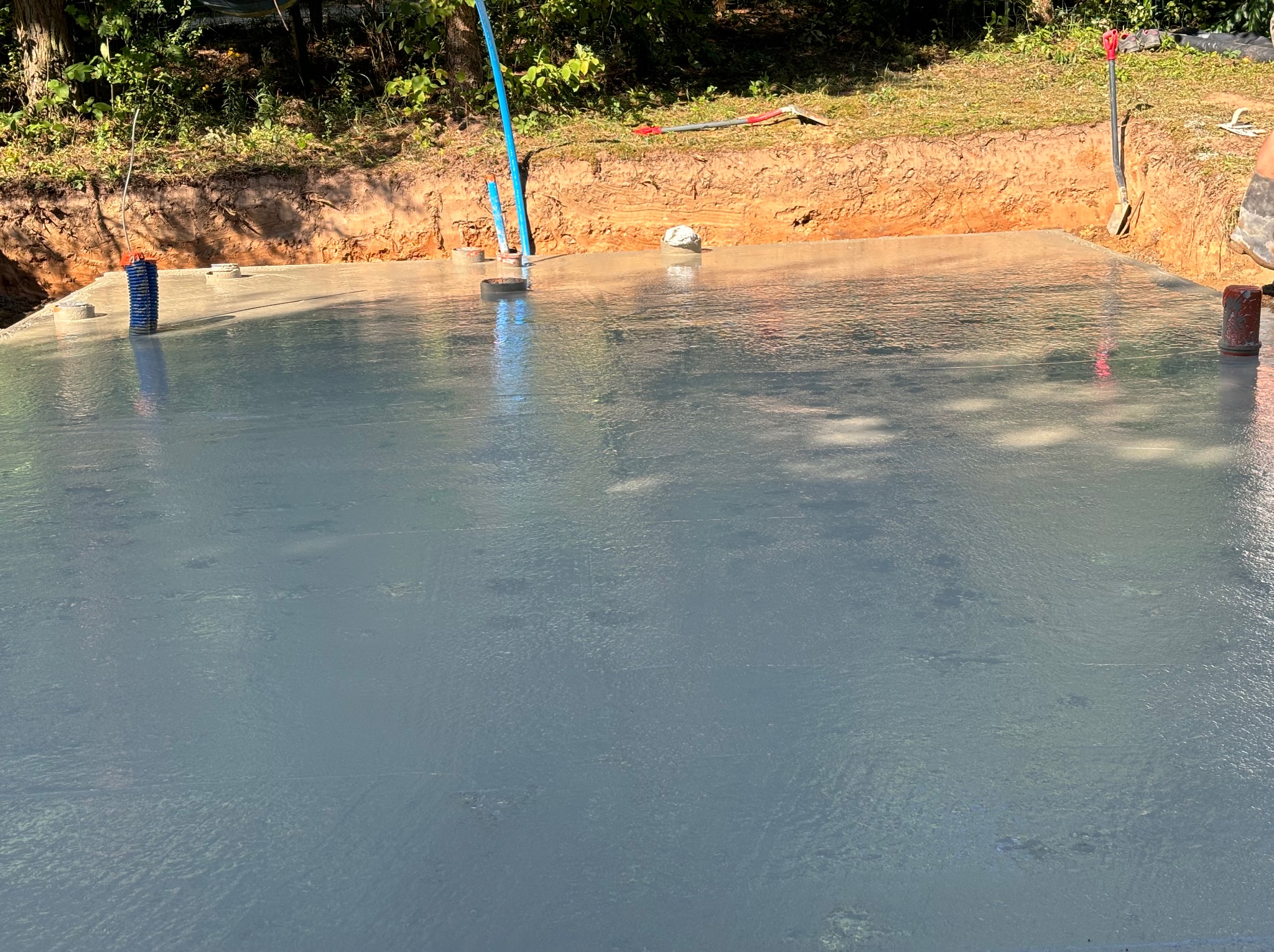 Świeżo wylana, gładka płyta fundamentowa z wystającymi rurami instalacyjnymi, otoczona wykopem i ziemią, widoczne narzędzia budowlane w tle.