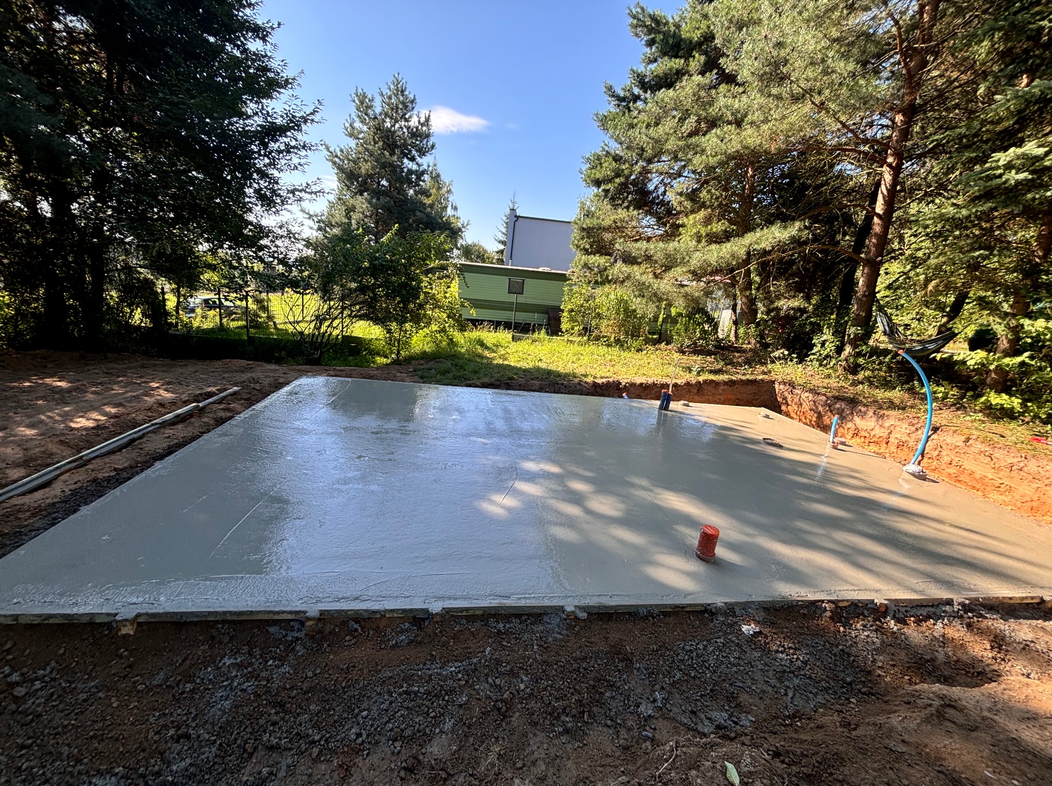 Świeżo wylana, mokra płyta betonowa fundamentowa z widocznymi elementami szalunku i wystającymi rurkami, otoczona ziemią i drzewami w słoneczny dzień.