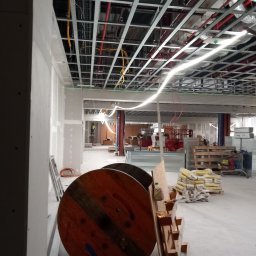 Dambud - Wnętrze w trakcie prac remontowych, widoczne szpachlowane ściany, metalowa konstrukcja sufitu podwieszanego z instalacjami, palety z workami zaprawy oraz bębny kablowe na pierwszym planie.