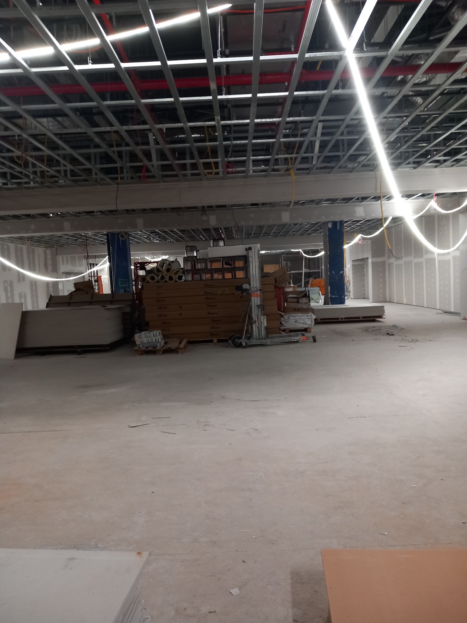 Wnętrze w trakcie prac wykończeniowych: widoczne szpachlowane ściany, metalowa konstrukcja sufitu podwieszanego, materiały budowlane składowane na podłodze i podnośnik platformowy.