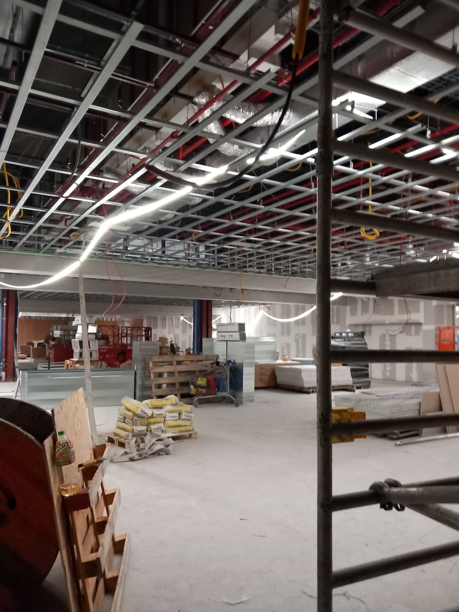 Wnętrze w trakcie prac wykończeniowych: widoczna metalowa konstrukcja sufitu podwieszanego, instalacje, materiały budowlane, rusztowanie i worki z zaprawą na posadzce.