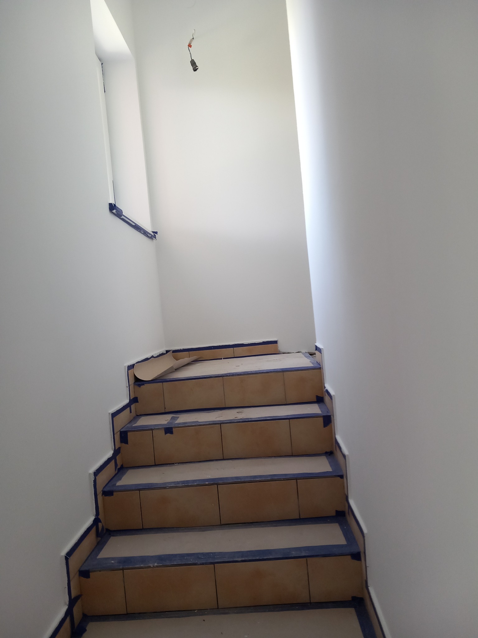 Schody wyłożone brązowymi płytkami, zabezpieczone niebieską taśmą malarską i kartonem, ściany pomalowane na biało, surowa żarówka zwisa z sufitu.