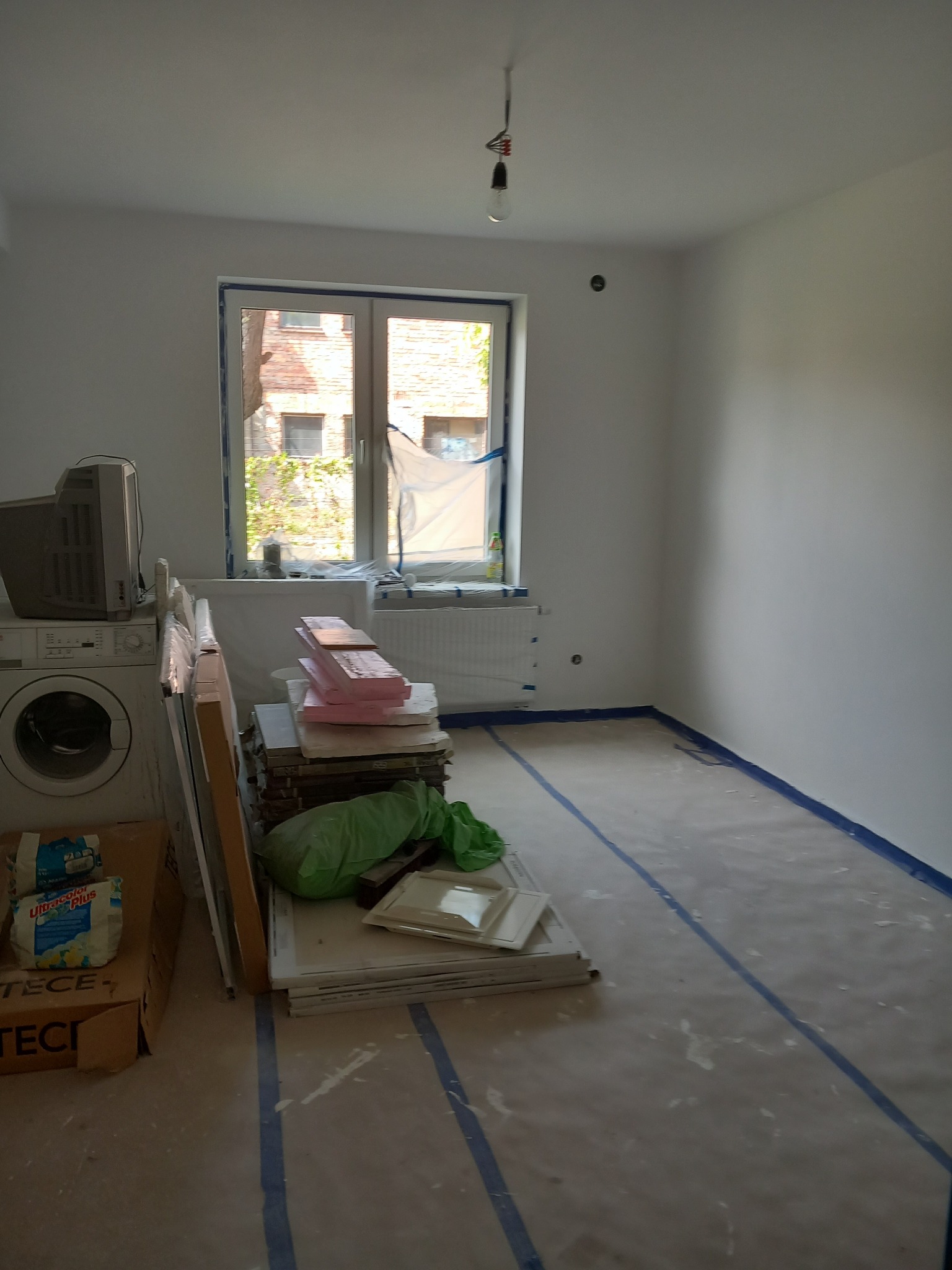 Pokój w trakcie remontu: zabezpieczona folią podłoga z taśmą malarską, materiały budowlane (płyty, styropian) przy ścianie, pralka i stary telewizor w tle, surowa żarówka na kablu zwisająca z sufitu.