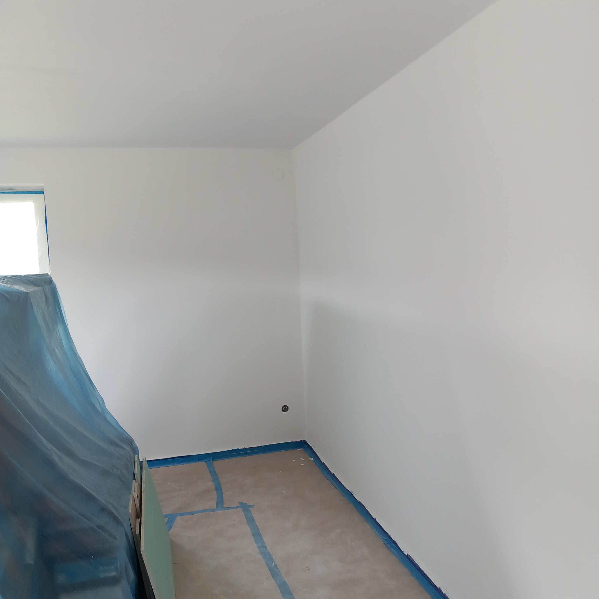 Pokój w trakcie prac przygotowawczych: świeżo pomalowane ściany na biało, zabezpieczona folią podłoga i okno, widoczne płyty gipsowe pod folią.