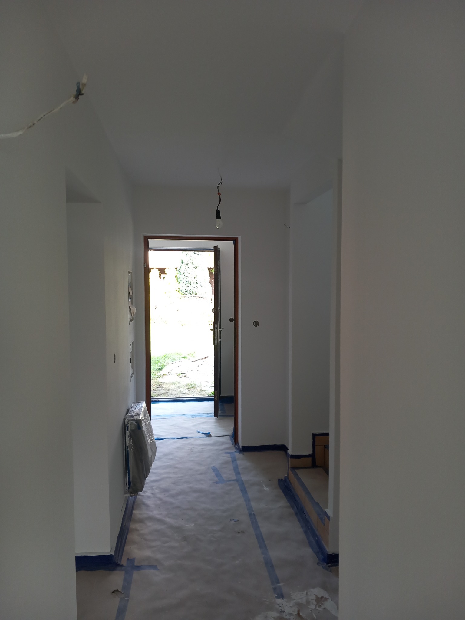 Korytarz w trakcie malowania na biało, podłoga zabezpieczona folią malarską z taśmą, widoczne niedociągnięcia w postaci zacieków na ścianie, w tle otwarte drzwi na zewnątrz.