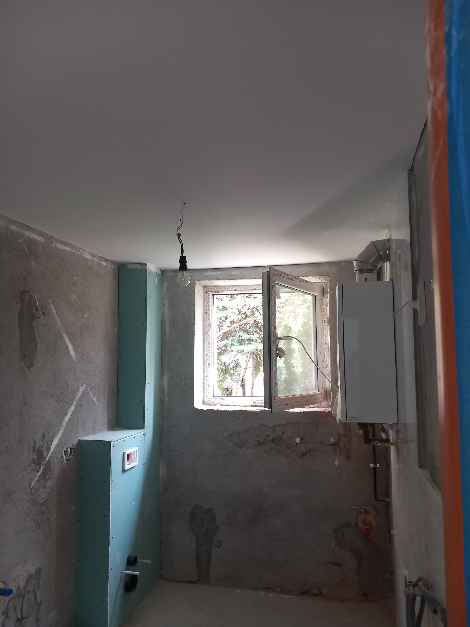 Surowa ściana w trakcie remontu łazienki z widoczną instalacją hydrauliczną, zabudową z płyt gipsowo-kartonowych, piecem gazowym i oknem z widokiem na zewnątrz.