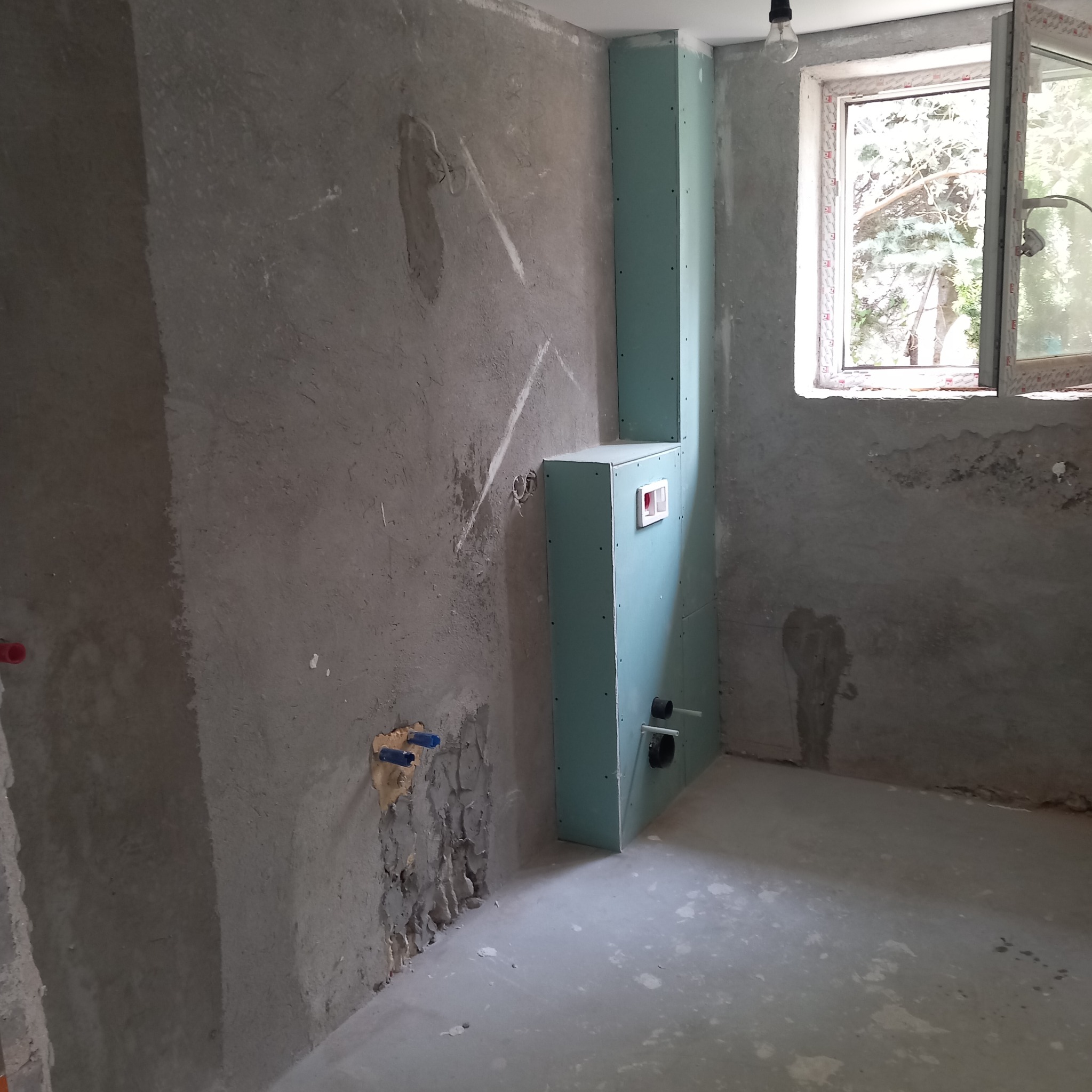 Surowa ściana z widocznymi instalacjami wodnymi i elektrycznymi, obudowana pionowa konstrukcja z płyt gipsowo-kartonowych z otworami na rury, przygotowanie do prac wykończeniowych, okno otwarte...