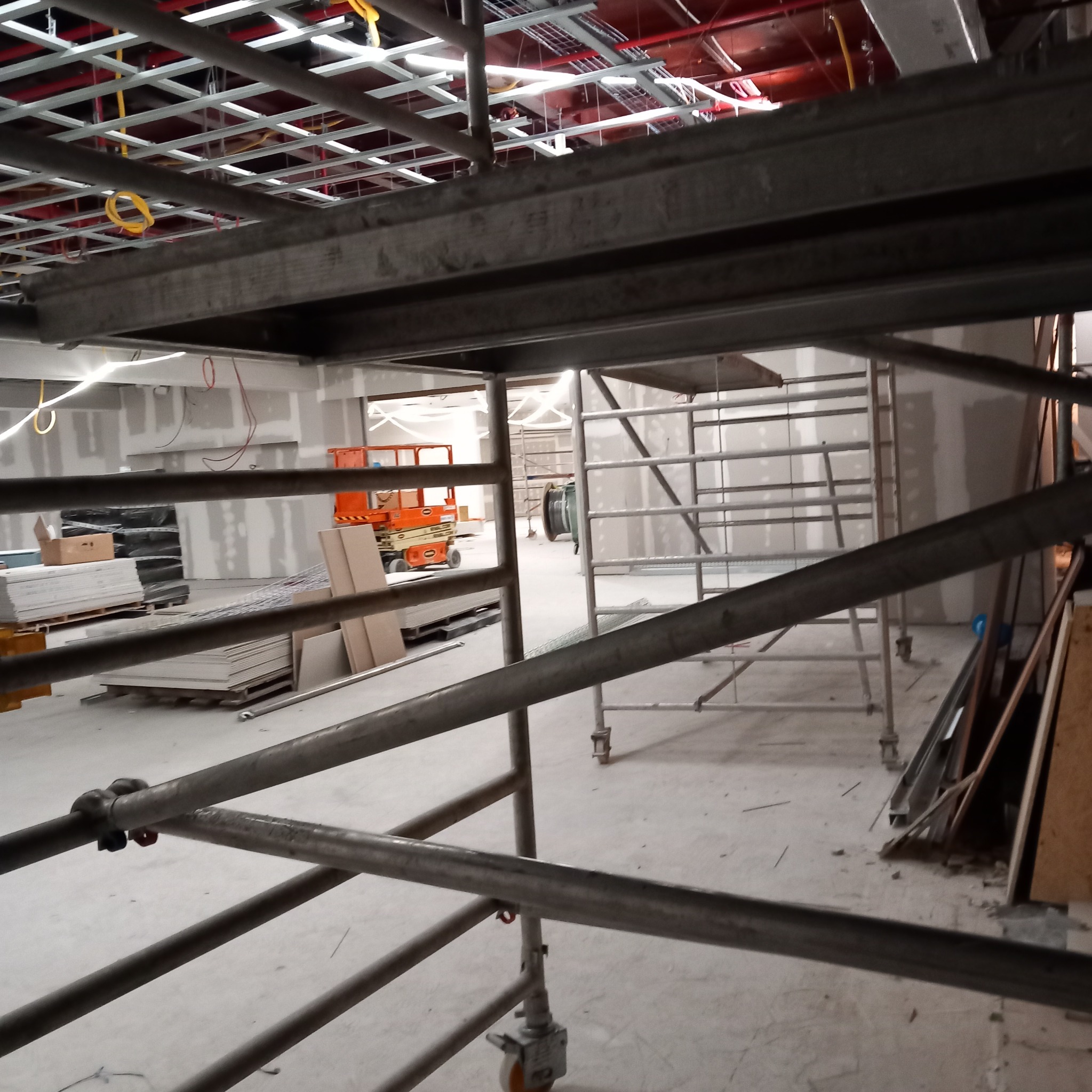 Wnętrze w trakcie prac wykończeniowych, widoczne rusztowania, płyty gipsowe i metalowa konstrukcja sufitu podwieszanego z instalacjami.
