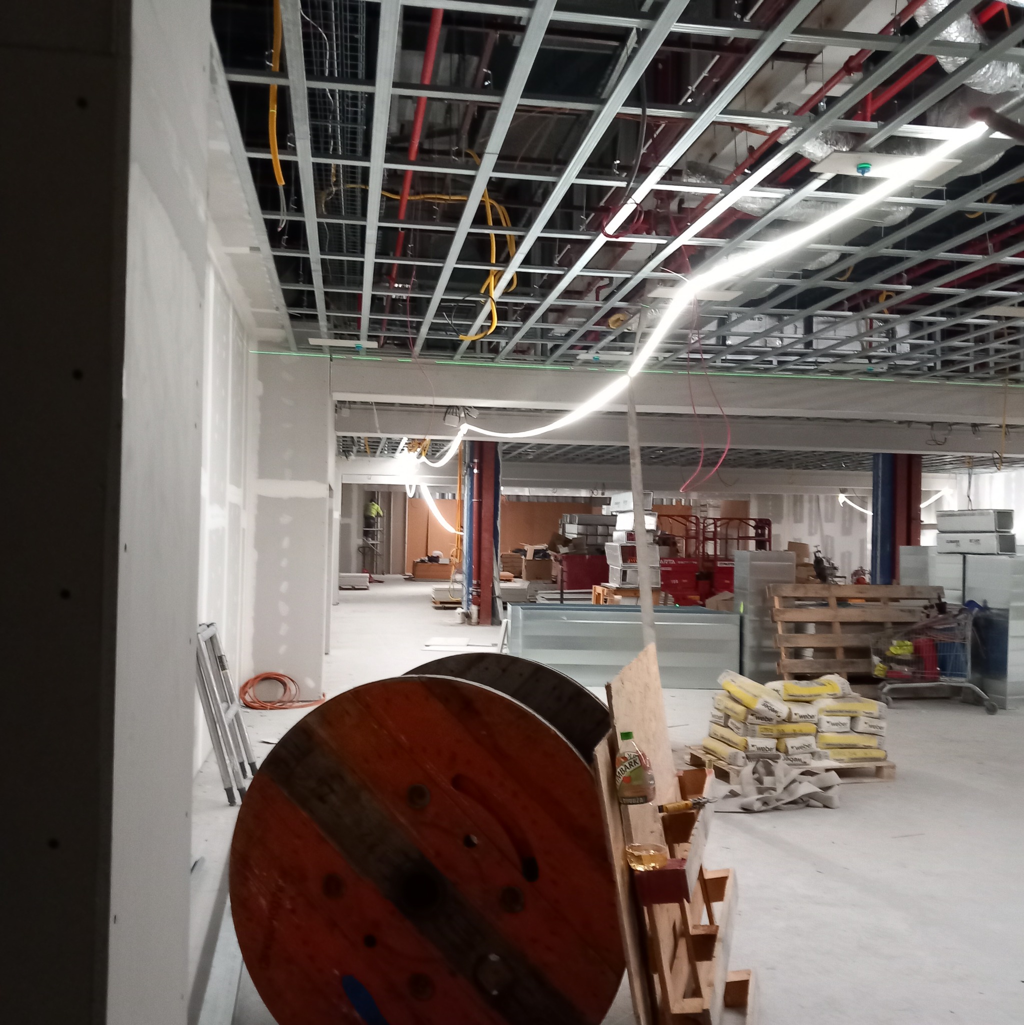 Wnętrze w trakcie prac remontowych, widoczne szpachlowane ściany, metalowa konstrukcja sufitu podwieszanego z instalacjami, palety z workami zaprawy oraz bębny kablowe na pierwszym planie.