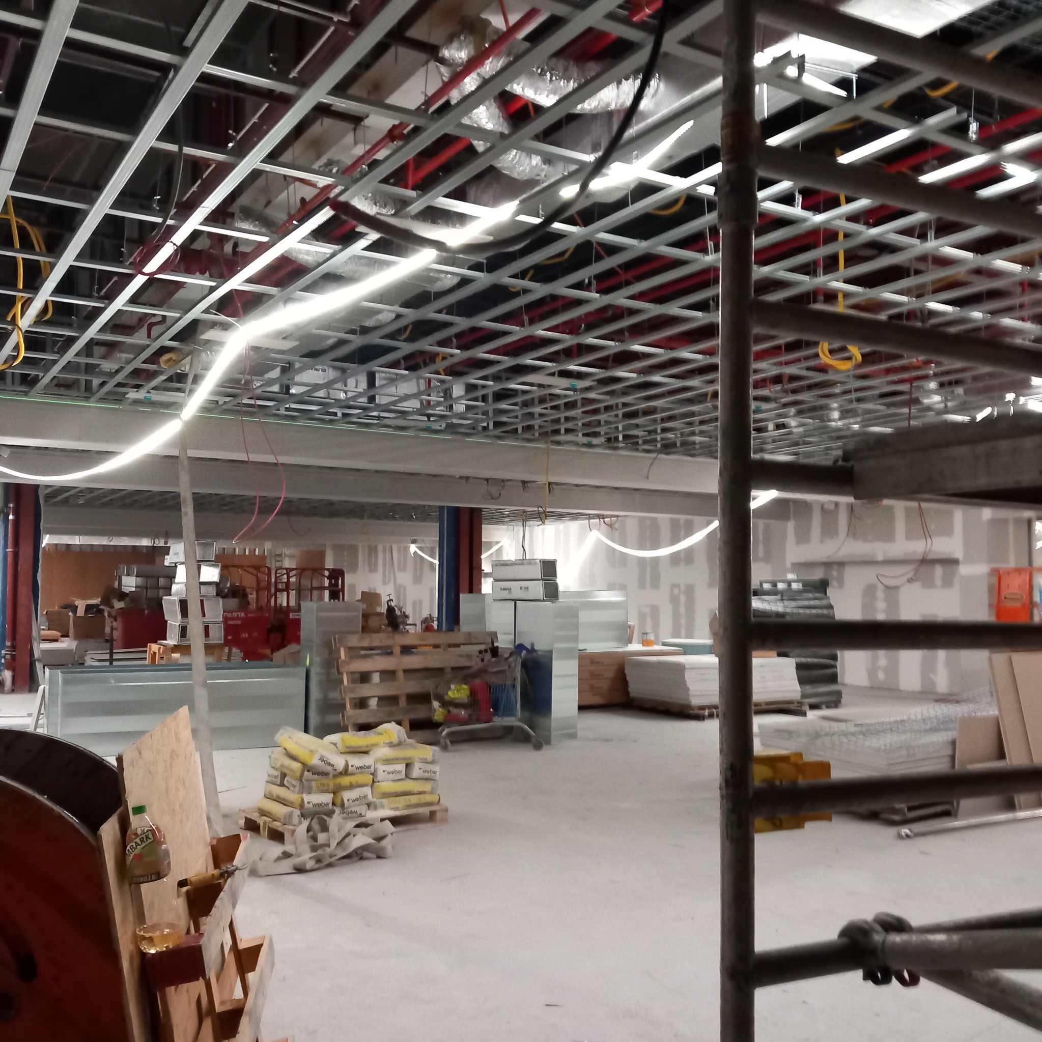 Wnętrze w trakcie wykończenia: widoczne metalowe profile sufitowe, instalacje, worki z zaprawą Weber na palecie, częściowo oszlifowane ściany, rusztowanie z przodu, butelka soku i szklanka...