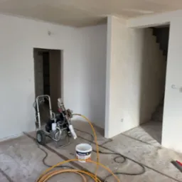 Pokój w trakcie prac wykończeniowych, widoczny agregat malarski, wiadro z farbą Dekorol i zabezpieczona podłoga folią malarską. Ściany świeżo pomalowane na biało.