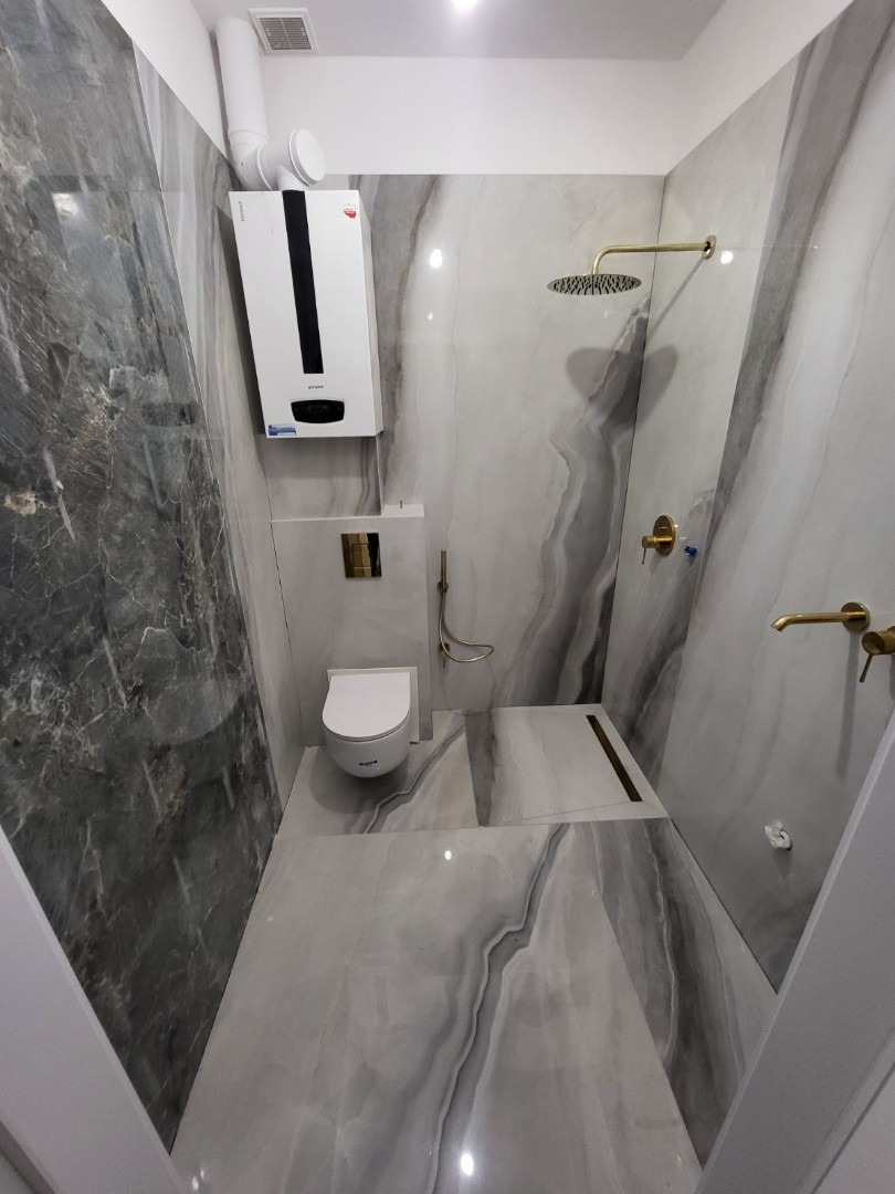 Wnętrze wykończonej łazienki z szarymi płytkami imitującymi marmur, złotą armaturą, podwieszaną toaletą i wiszącym bojlerem.
