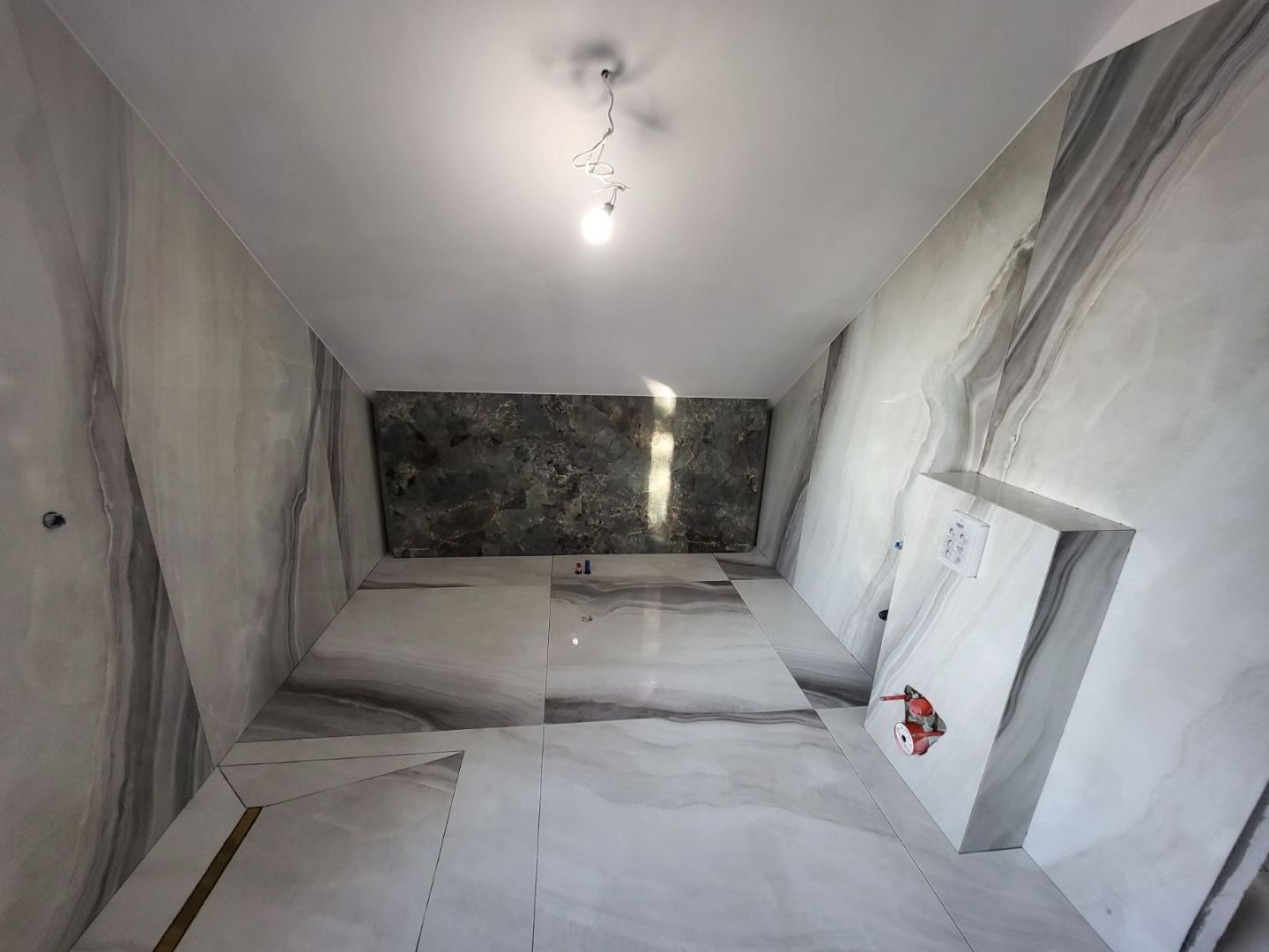Wnętrze łazienki w trakcie wykańczania, z marmurkowymi płytkami na ścianach i podłodze, widoczne przyłącza wodne i elektryczne oraz fragment podwieszanego stelaża WC.
