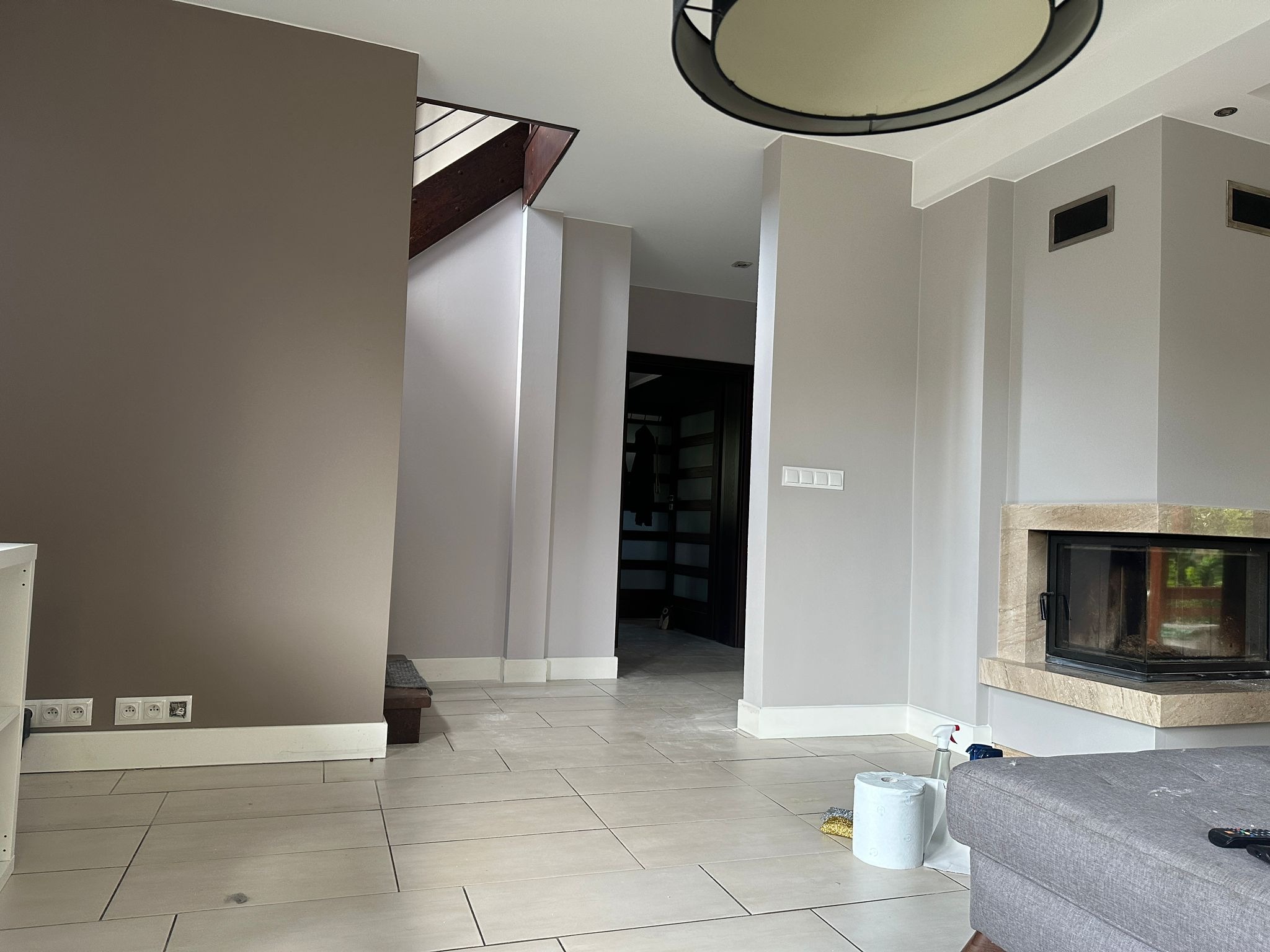 Wnętrze po malowaniu, widoczna ściana w odcieniach brązu, szare płytki podłogowe, kominek z kamienną obudową i fragment sofy, gniazdka elektryczne przy podłodze.