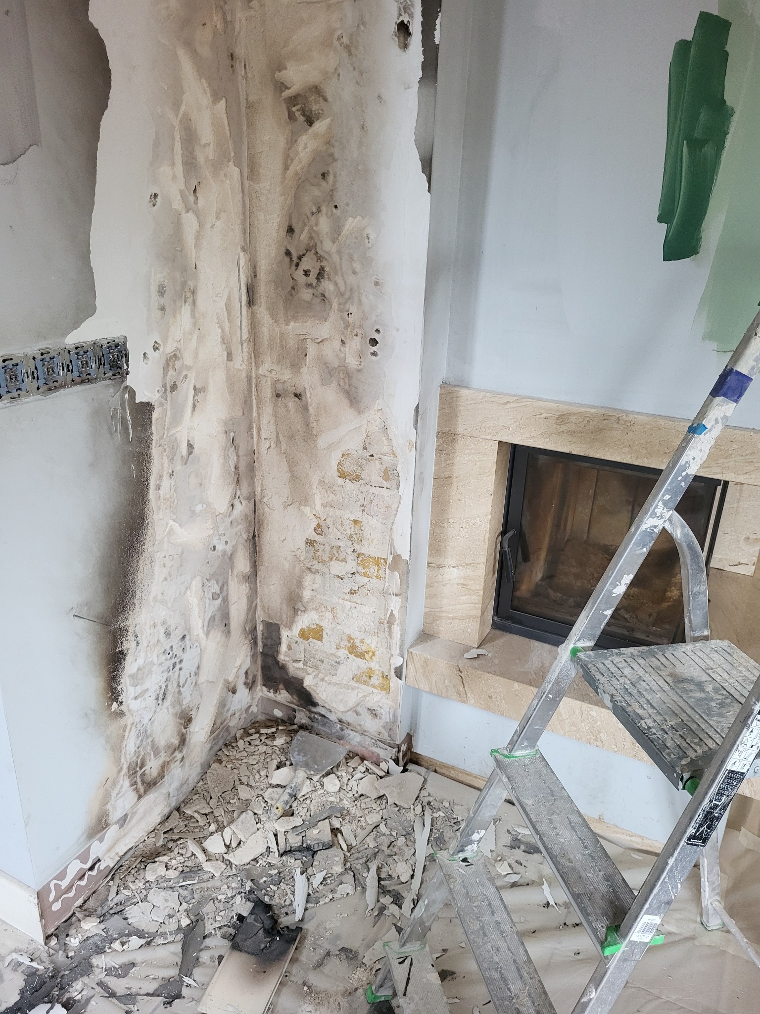 Ściana w trakcie demontażu starej okładziny, widoczne fragmenty cegły i resztki tynku, drabina oparta o ścianę z kominkiem, przygotowania do prac remontowych.