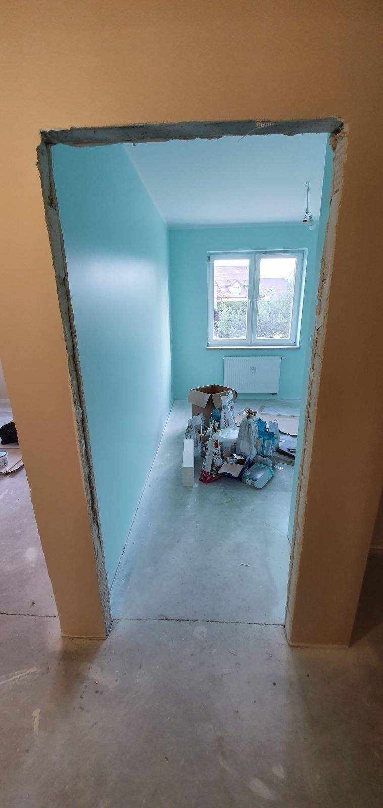 Wnętrze pokoju z pomalowanymi na błękitno ścianami, widoczne przez surowy otwór drzwiowy, na podłodze karton i worki z materiałami budowlanymi.