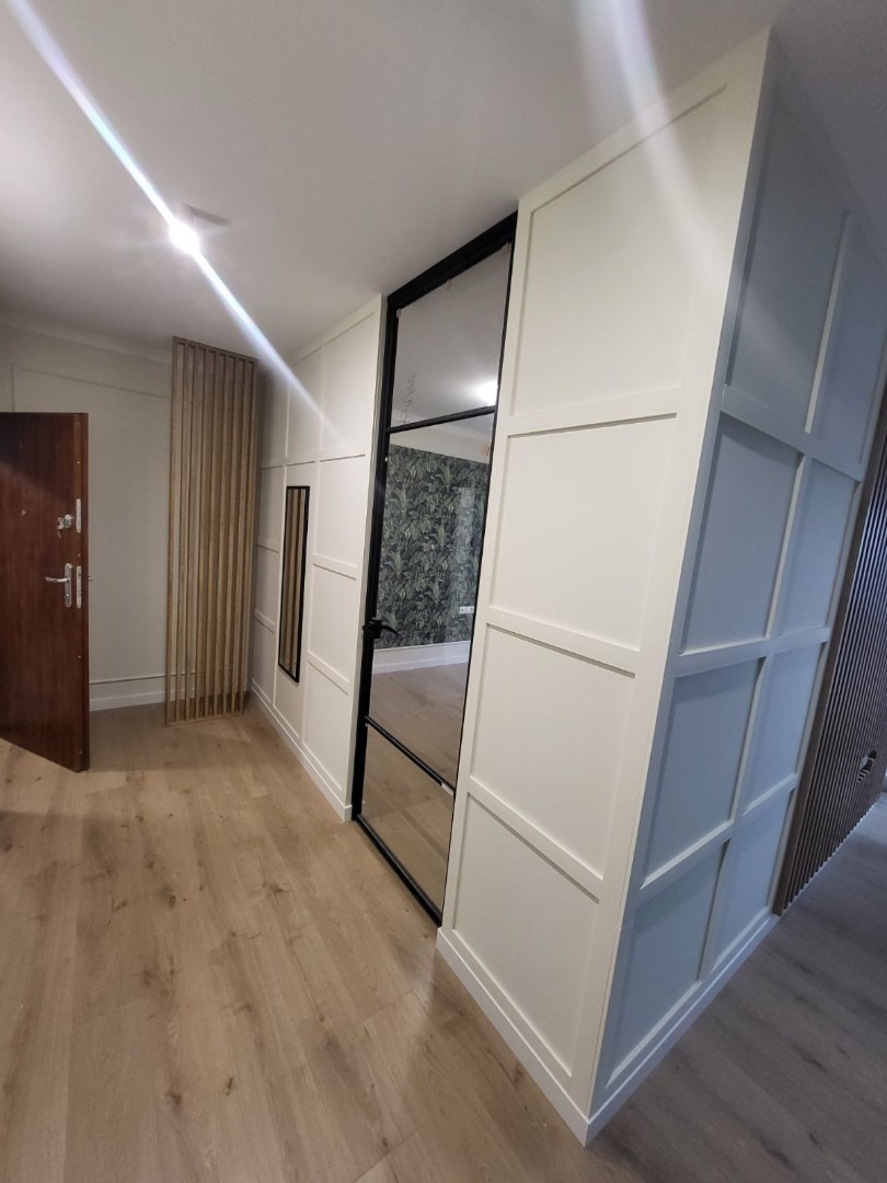 Nowoczesne wykończenie przedpokoju w Gdyni: białe panele ścienne z czarnymi akcentami, drewniana podłoga i ażurowa ścianka działowa z drewna.