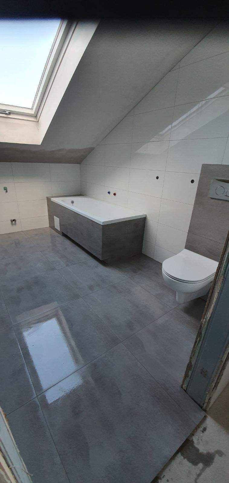 Nowoczesna łazienka na poddaszu z oknem dachowym, szare płytki podłogowe z połyskiem, białe płytki ścienne, zabudowana wanna w szarej obudowie i podwieszana toaleta.