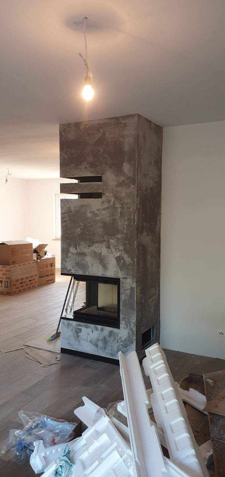 Surowy kominek z gładzią dekoracyjną w trakcie wykańczania, widoczny fragment podłogi z panelami i resztki materiałów budowlanych, surowa żarówka na kablu.