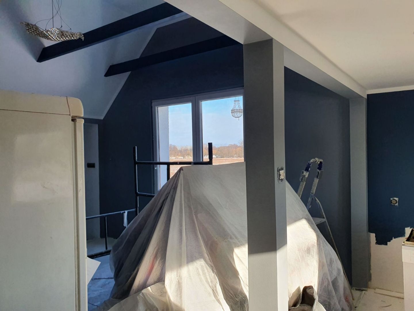 Pokój w trakcie prac wykończeniowych: ściany w odcieniach granatu i szarości, zabezpieczone meble pod folią, drabina malarska, widoczne elementy oświetlenia.