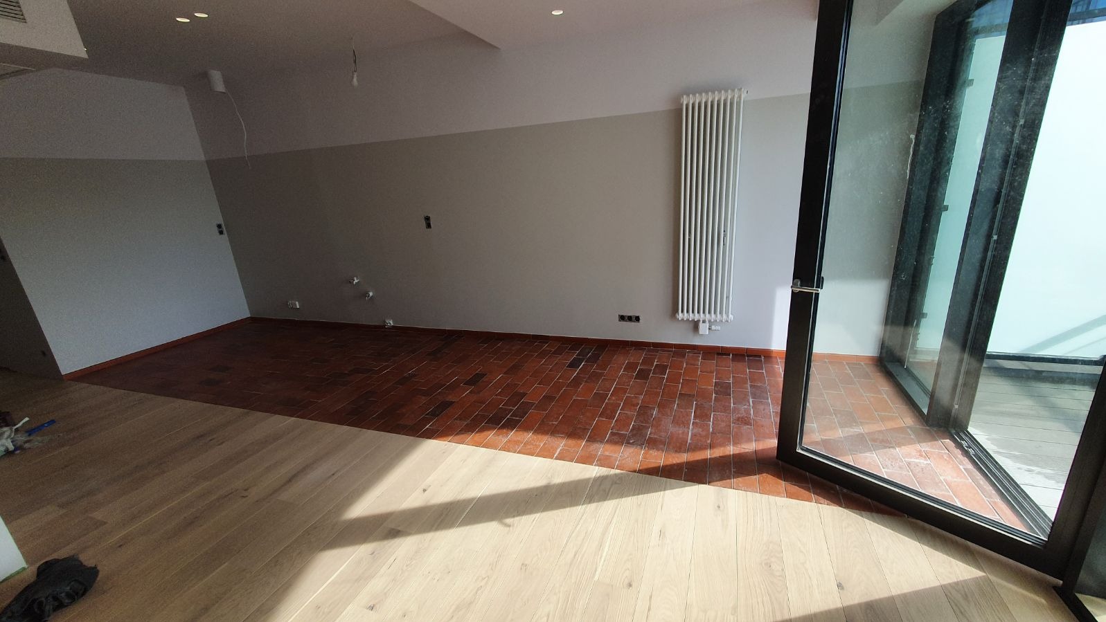 Pokój z częściowo ułożoną podłogą: połączenie jasnych paneli i czerwonej cegły, ściany pomalowane na szaro-biało, duży grzejnik i otwarte, nowoczesne okno balkonowe.