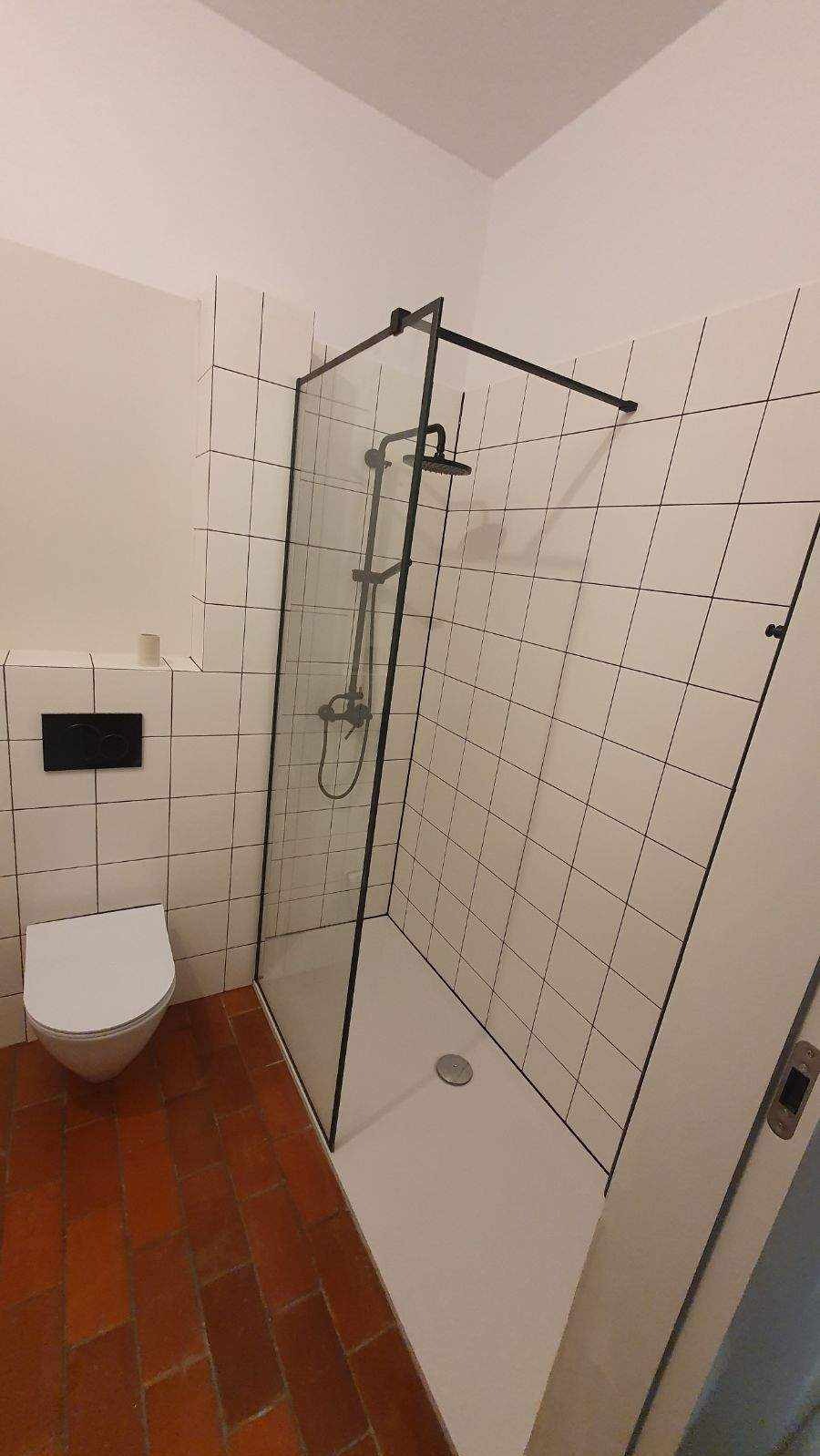 Nowoczesna kabina prysznicowa z czarną ramą i deszczownicą, białe kwadratowe płytki z czarną fugą, terakota na podłodze, wisząca toaleta z czarnym przyciskiem.