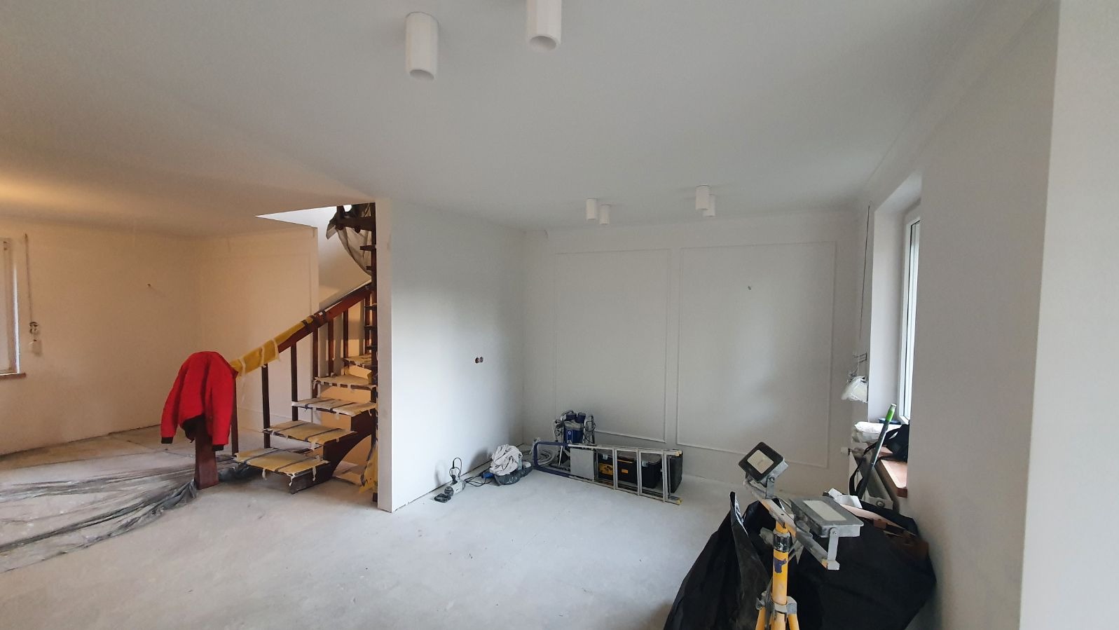 Wnętrze w trakcie prac wykończeniowych: świeżo zagruntowane ściany, zabezpieczone schody z drewnianą balustradą, drabina i sprzęt oświetleniowy w centralnej części pomieszczenia, widoczne gniazdka...