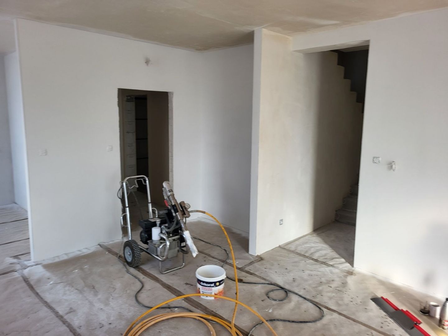 Pokój w trakcie prac wykończeniowych, widoczny agregat malarski, wiadro z farbą Dekorol i zabezpieczona podłoga folią malarską. Ściany świeżo pomalowane na biało.