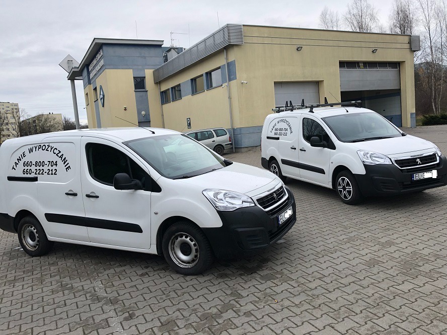 Dwa białe samochody dostawcze marki Peugeot zaparkowane na kostce brukowej przed budynkiem. Na boku jednego z aut widnieje napis 'Tanie Wypożyczanie' oraz numery telefonów.