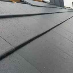 Nowy, grafitowy dach z dachówki ceramicznej, pokryty kroplami deszczu, z widoczną więźbą dachową i kominem w tle.