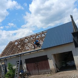 MassDach - Pracownik w niebieskiej koszulce montuje blachodachówkę na drewnianej konstrukcji dachu budynku gospodarczego, widoczne drabiny i narzędzia na zewnątrz.
