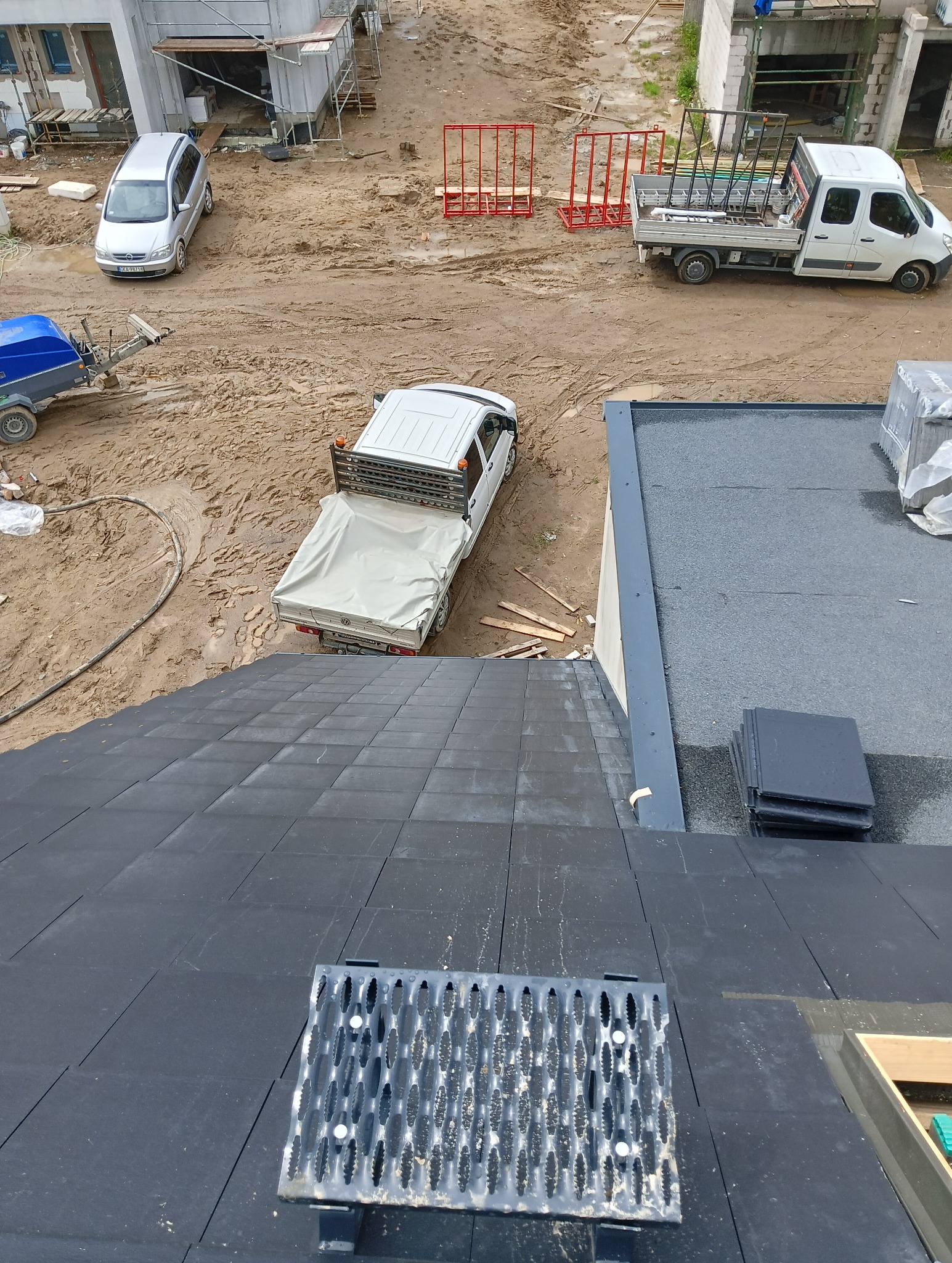 Widok z góry na nowo ułożony, czarny dach z płytkami, z metalową kratką ułatwiającą poruszanie się po dachu; w tle plac budowy z zaparkowanymi samochodami i materiałami budowlanymi.