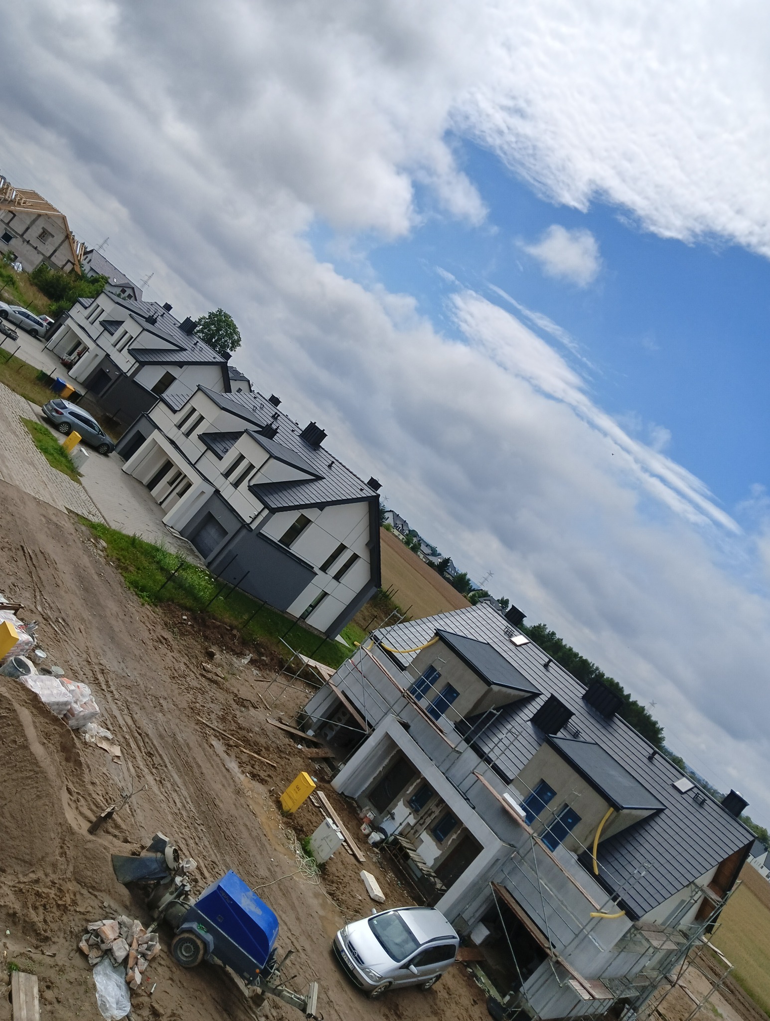 Nowe domy w zabudowie szeregowej z ciemnymi dachami, niektóre w trakcie budowy, widoczne rusztowania i sprzęt budowlany, pochmurne niebo.