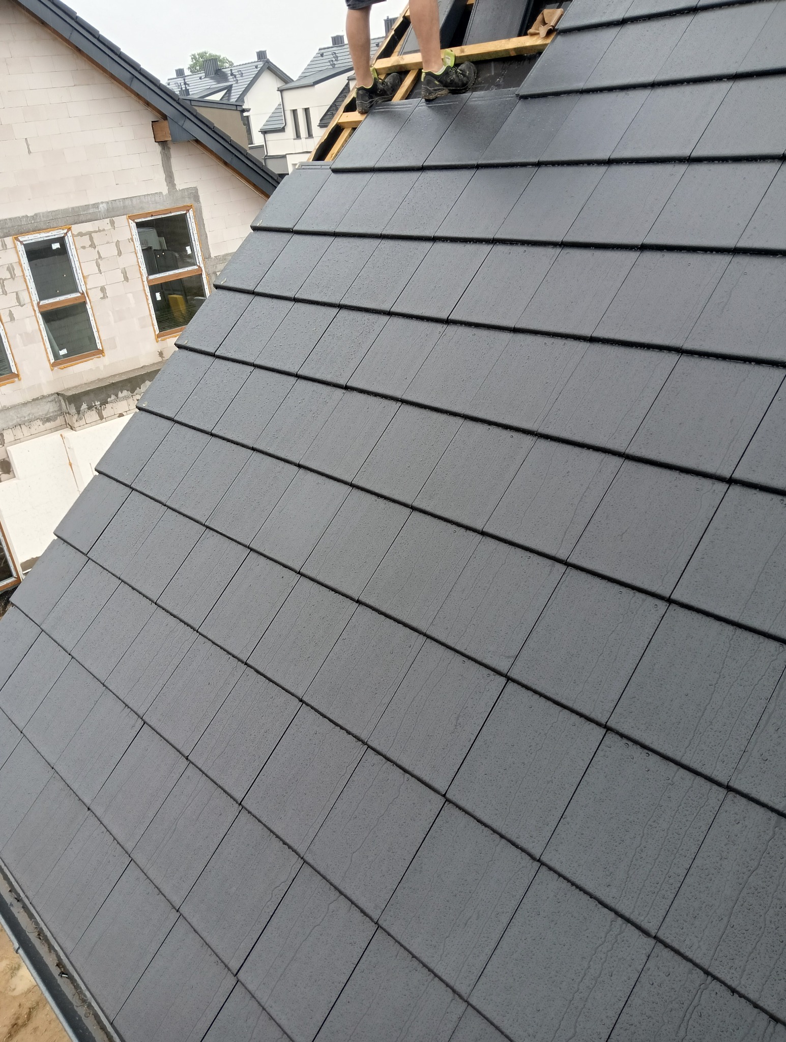 Nowo położona, grafitowa dachówka ceramiczna na skośnym dachu domu w trakcie budowy, widoczne fragmenty więźby i stopy dekarza.