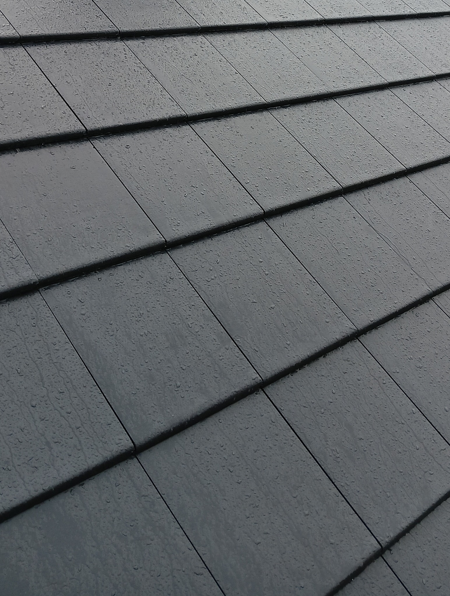 Szczegółowe ujęcie czarnych dachówek pokrytych kroplami deszczu, ułożonych ukośnie, z widoczną fakturą i precyzyjnym łączeniem.