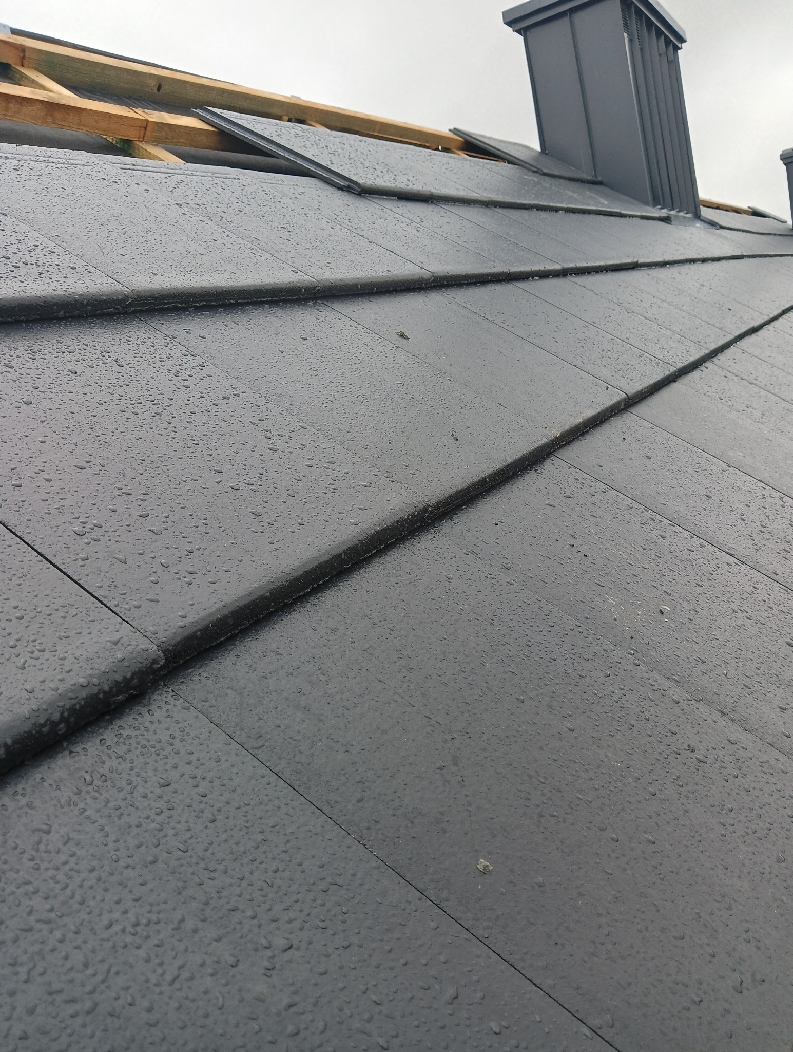 Nowy, grafitowy dach z dachówki ceramicznej, pokryty kroplami deszczu, z widoczną więźbą dachową i kominem w tle.