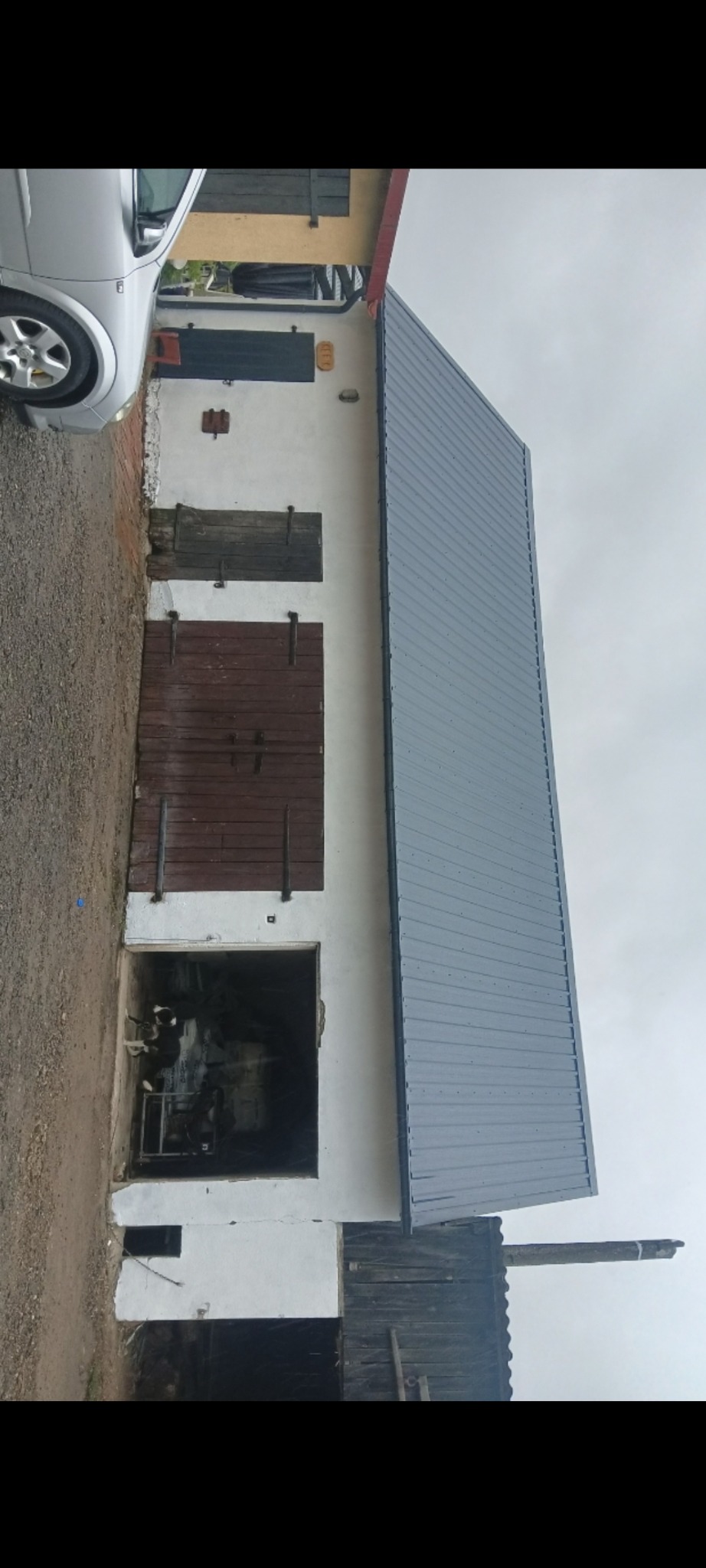 Metalowy dach w kolorze szarym na budynku gospodarczym z białymi ścianami i drewnianymi wrotami, widoczny fragment srebrnego samochodu