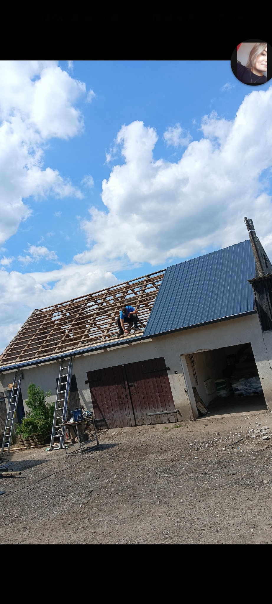 Pracownik w niebieskiej koszulce montuje blachodachówkę na drewnianej konstrukcji dachu budynku gospodarczego, widoczne drabiny i narzędzia na zewnątrz.