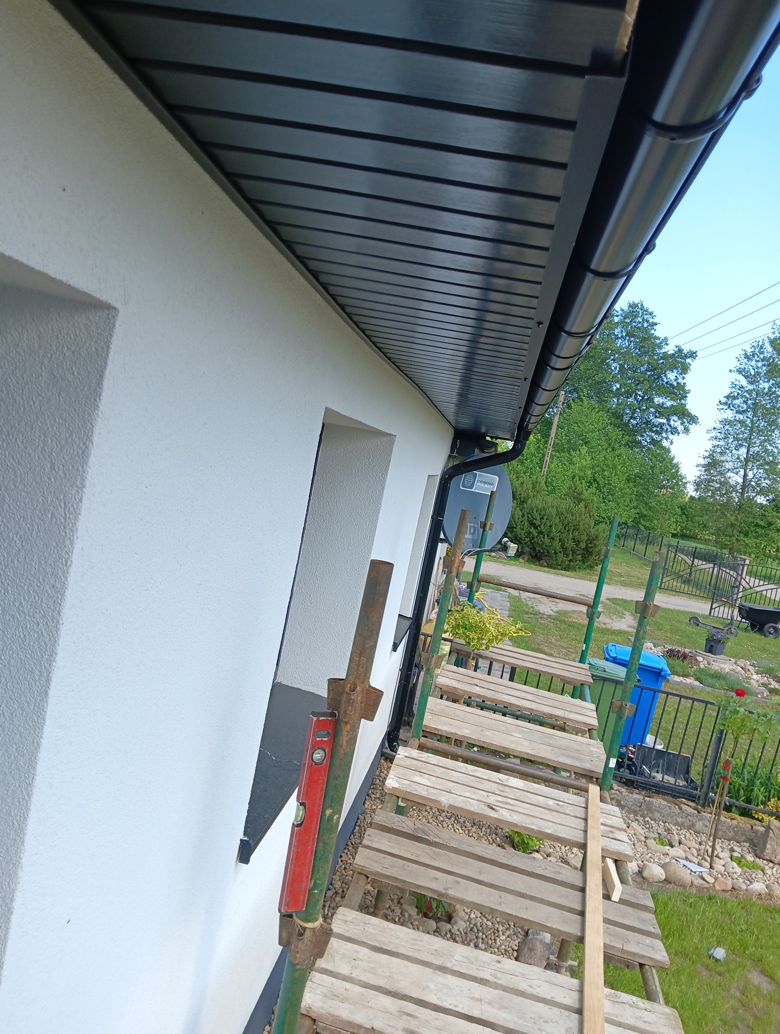 Fragment domu z nową, czarną podbitką dachową i rynną, widoczne z poziomu rusztowania z poziomicą przymocowaną do rury.