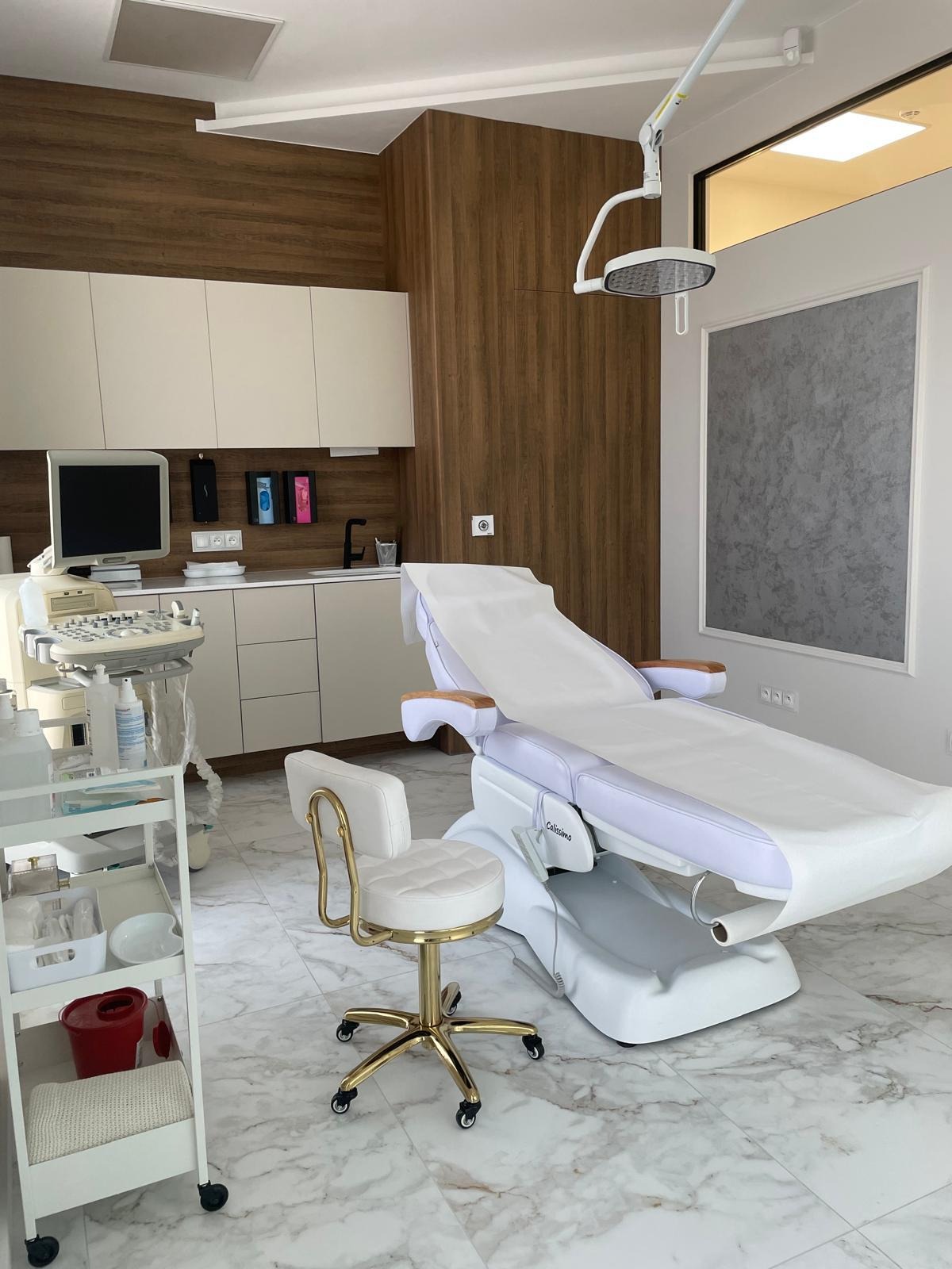 Nowoczesne wnętrze gabinetu kosmetycznego z białym fotelem zabiegowym, złotym krzesłem, szafkami w odcieniach beżu i marmurową podłogą. Widoczny sprzęt medyczny i oświetlenie punktowe.