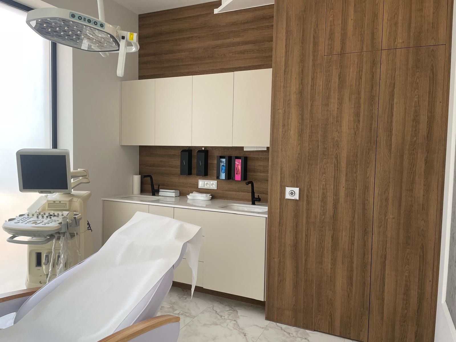 Nowoczesne wnętrze gabinetu lekarskiego z drewnianymi akcentami, białymi szafkami, umywalkami z czarnymi bateriami, aparatem USG i fotelem pacjenta.