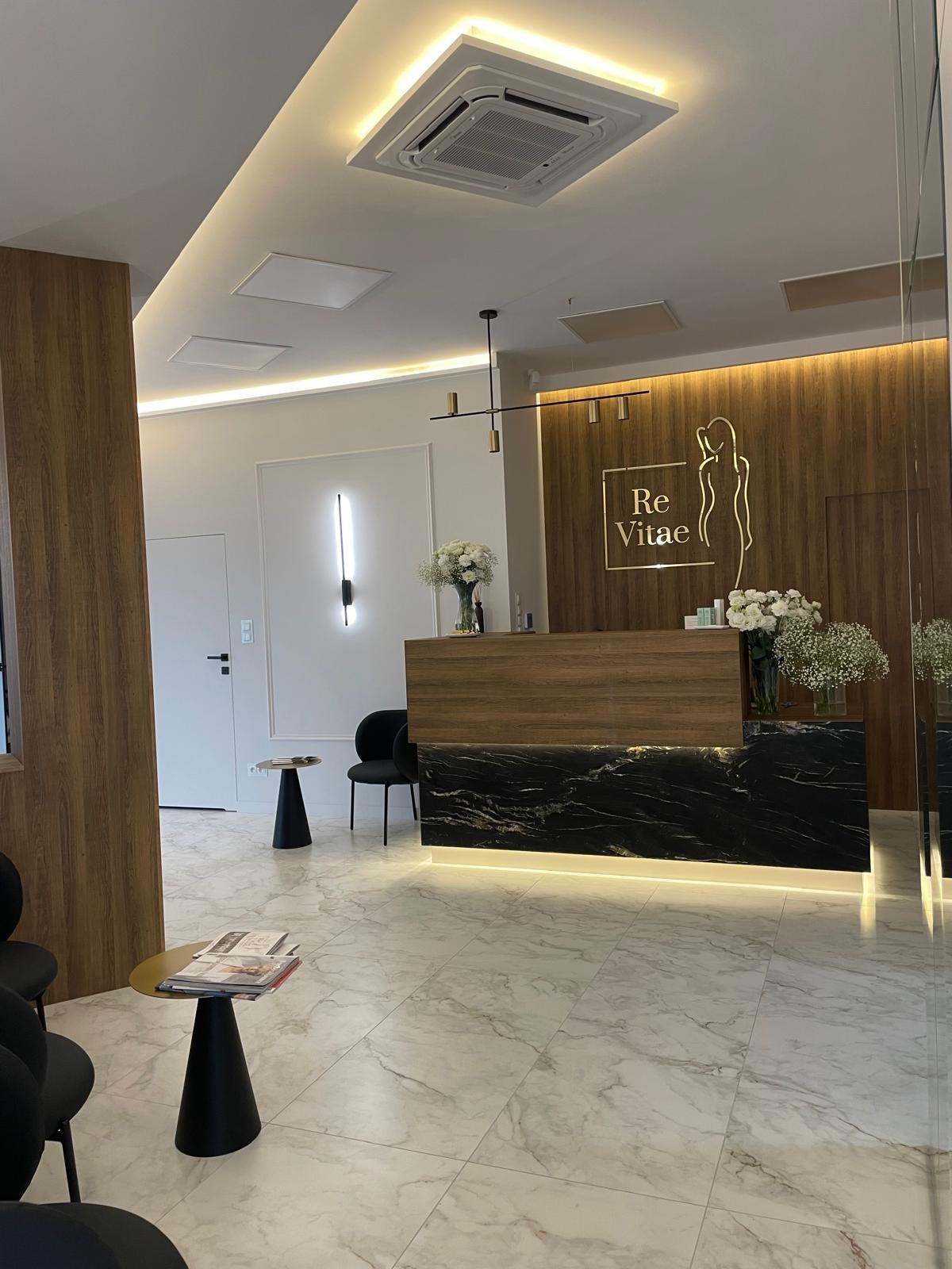 Nowoczesna recepcja z drewnianą ścianą, marmurowym blatem i podświetleniem LED, minimalistyczna lampa sufitowa, biała marmurowa podłoga i czarne krzesła w tle.