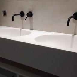 Ergonomic Marta Szponarska - Podwójna, biała umywalka z czarnymi, ściennymi bateriami w nowoczesnej łazience z surowym wykończeniem ścian.