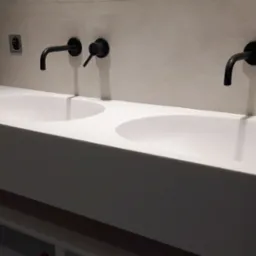 Podwójna, biała umywalka z czarnymi, ściennymi bateriami w nowoczesnej łazience z surowym wykończeniem ścian.