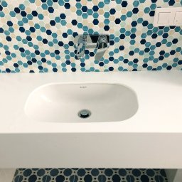 Ergonomic Marta Szponarska - Biała, prostokątna umywalka z chromowaną baterią na tle mozaiki z heksagonów w odcieniach błękitu i bieli, z widocznym fragmentem toalety z identyczną mozaiką na podłodze.