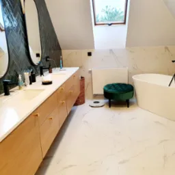 Nowoczesna łazienka na poddaszu z długim drewnianym blatem, owalnymi lustrami, zieloną mozaiką na ścianie i wolnostojącą białą wanną.