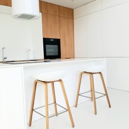 Ergonomic Marta Szponarska - Minimalistyczna kuchnia z białym blatem, drewnianymi szafkami i dwoma hokerami z drewnianymi nogami i białymi siedziskami, oświetlona naturalnym światłem.