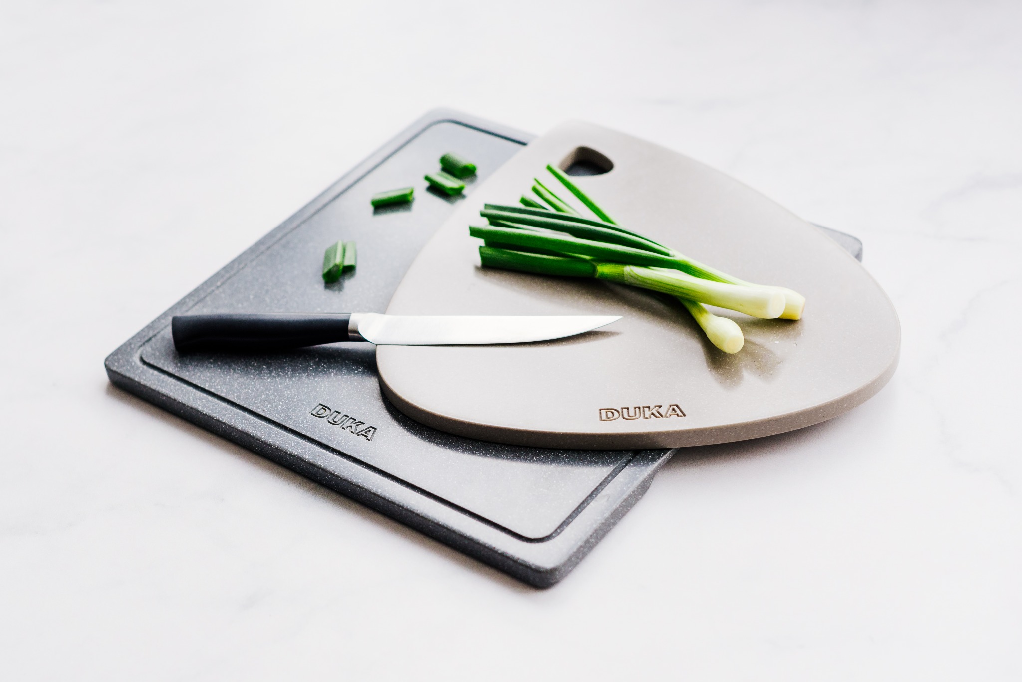 Dwie deski do krojenia marki DUKA: szara kwadratowa i beżowa o zaokrąglonym kształcie, z nożem i posiekanym szczypiorkiem na marmurowym blacie.